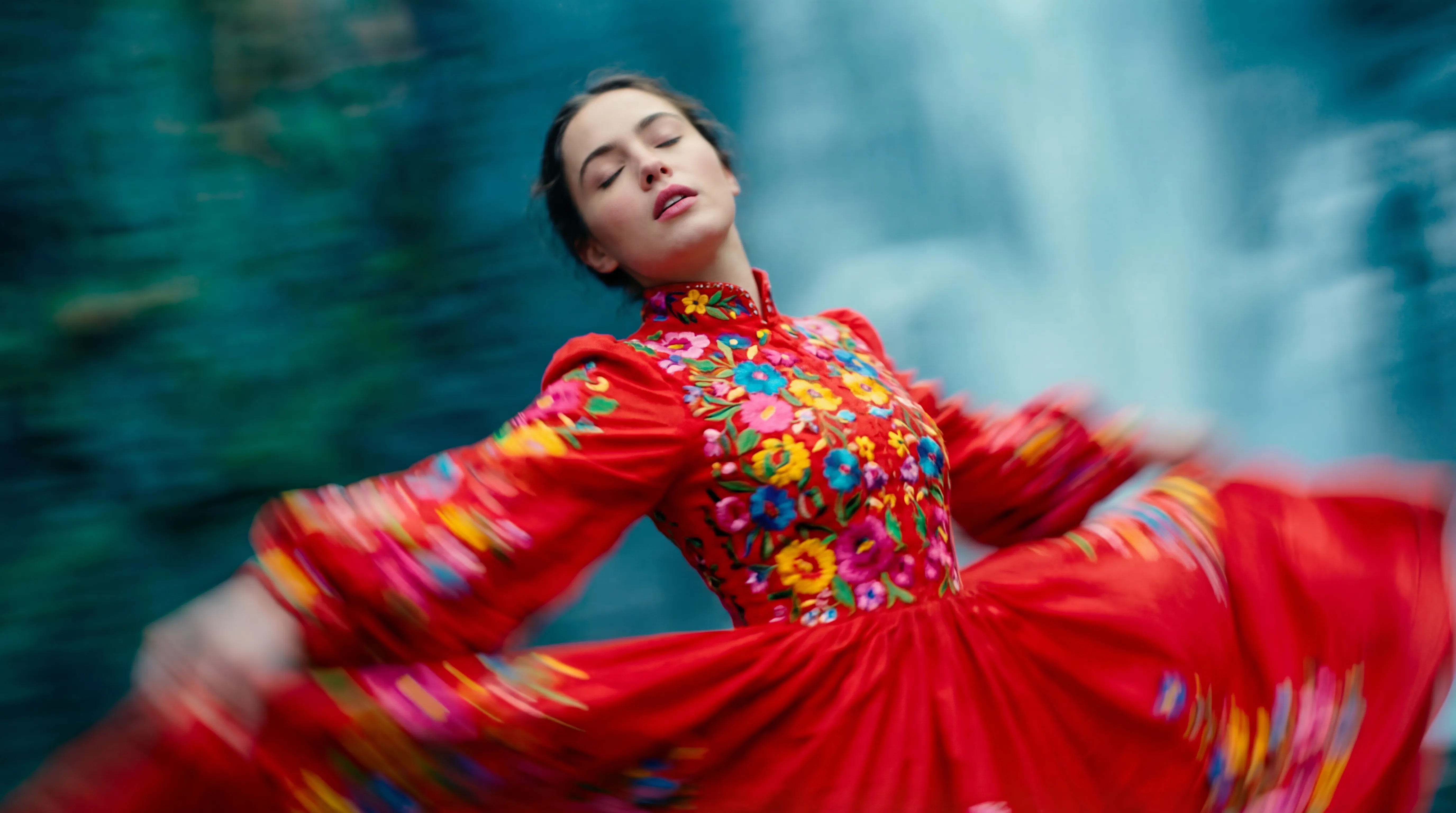 This cinematic still, in the style of a vibrant, avant-garde art film, captures a dynamic moment reminiscent of a bold, experimental visual storyteller's work. The camera is angled from a low perspective, looking upwards at a young woman caught in a blurred motion, giving the impression of exhilarating speed or a sudden, graceful turn. Her eyes are closed, head slightly tilted back, and lips softly parted, suggesting a moment of profound immersion or a dramatic exhalation. She is adorned in a striking, voluminous gown, predominantly a rich, saturated red, with the bodice and high collar intricately embellished with a riot of intensely colorful floral embroideryâhues of yellow, blue, green, and pink exploding against the fiery red. The fabric of her dress, especially the sleeves and lower skirt, is dramatically streaked and blurred by the motion, creating a sense of fluidity and energy that radiates outwards from her form. The background is an abstract blur of cool tones, with swathes of deep teal and lighter, almost ethereal blues, suggesting an environment that is fast-moving or out of focus, further emphasizing the woman as the sharp, albeit moving, subject. The overall ambiance is one of intoxicating freedom, passionate expression, and dreamlike exhilaration, underscored by the blurred edges and intense color palette, conveying a breathtaking, ephemeral instant.
