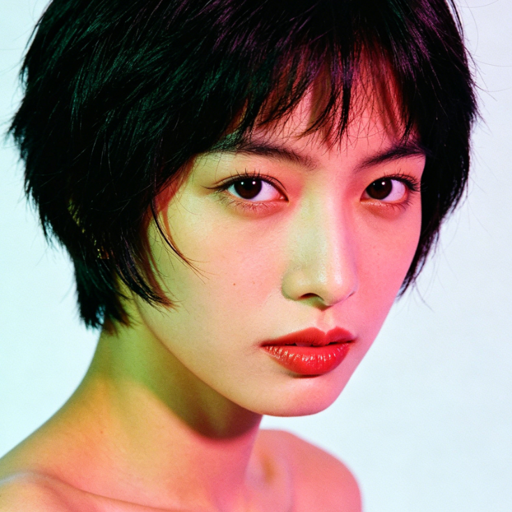 Portrait close-up of a woman with an AI 1980s filter applied. She has short black hair with layered ends and natural bangs. Her skin displays a soft pink-green gradient tone due to the filter, with bright red lipstick on her lips. She looks directly at the camera, creating a retro and artistic atmosphere.