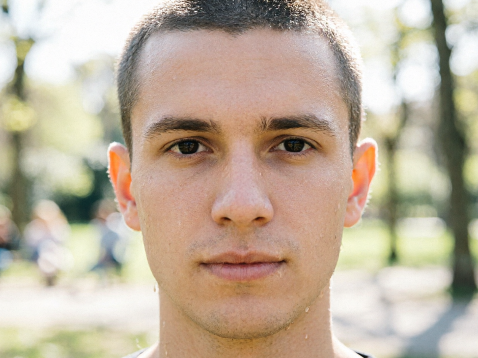 Frontal portrait of a young European man in outdoor sunlight, short hair with natural dark irises, clean-shaven face, everyday realistic style, 4:3 aspect ratio