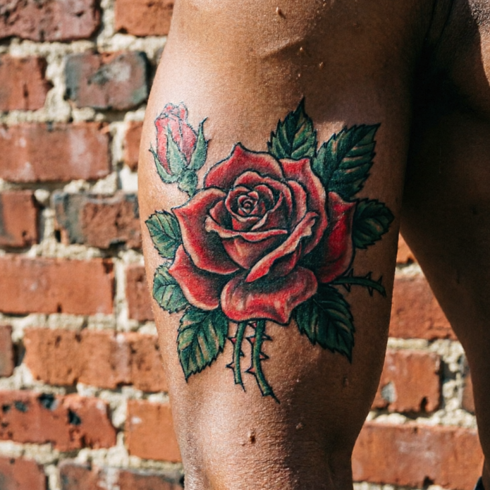 Full-bloom red rose tattooed on outer thigh, layered petals with serrated green leaves and thorny stem; unopened bud above against red brick wall.