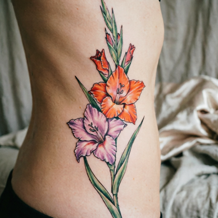 Two gladiolus flowers tattooed on a woman’s side waist—one orange-red, one pink-purple—with layered petals and slender green leaves; vibrant colors against soft fabric and indoor setting.