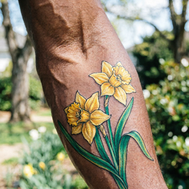 Two yellow daffodils tattooed on dark-skinned arm, full petals with trumpet-shaped centers and long green leaves; crisp lines against outdoor greenery and sunlight.