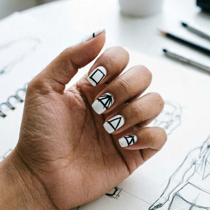 Hand on sketchbook; nails in pure white with black geometric shapes like squares, triangles, and arrows; pencils and sketches in background reflect modern minimalist artistry.