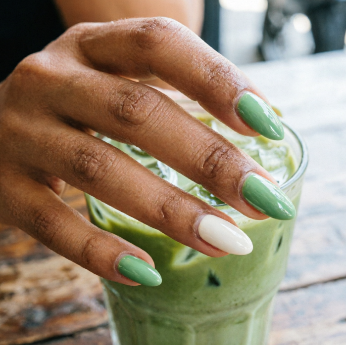 Hand holding iced matcha latte; mint green nails with one pure white accent nail create fresh contrast against wooden table backdrop.