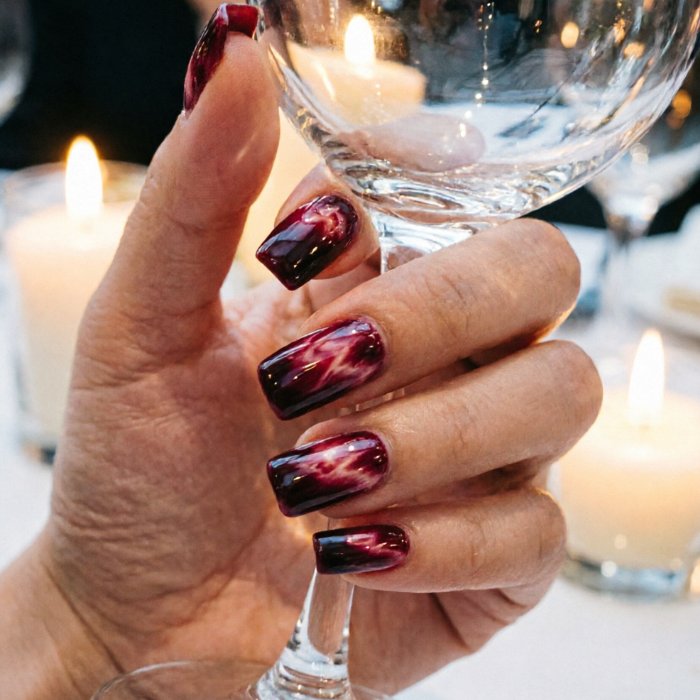 Deep burgundy nails with white marble pattern grip a wine glass; candlelit dinner background enhances luxurious romance.
