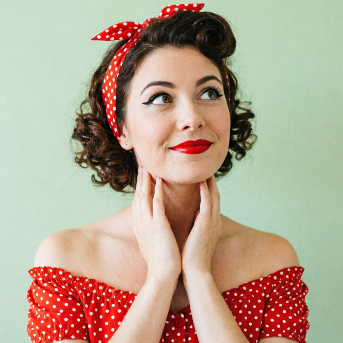 A portrait of a young woman wearing a red and white polka-dot off-the-shoulder top and matching headband with retro curls. She has bright red lipstick, cat-eye makeup, and is resting her chin on her hands while looking upwards playfully against a mint green background.