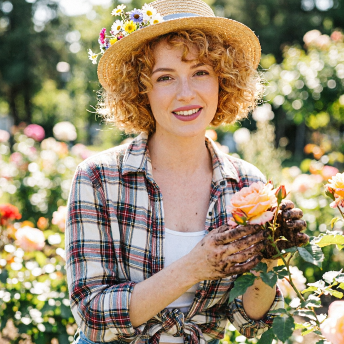 A curly-haired woman in a flower-adorned straw hat and plaid shirt stands in a rose garden, gently holding a pink rose with soil-stained hands, smiling softly amidst blooming flowers to evoke serene countryside vibes.