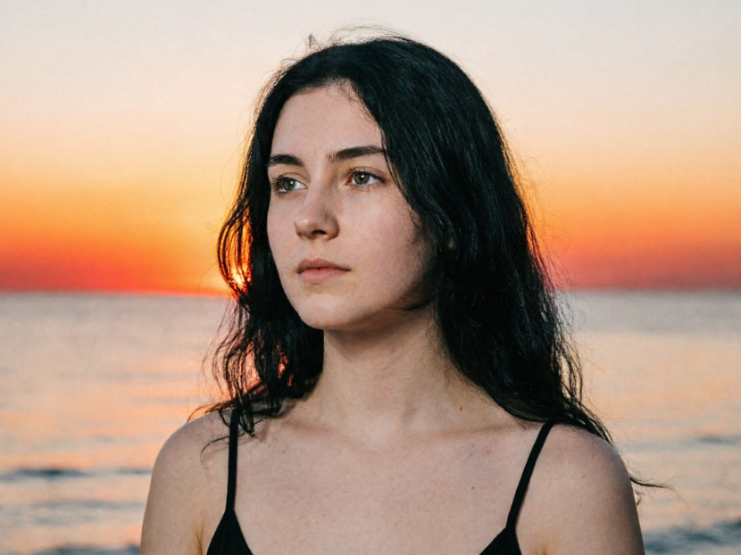 An outdoor portrait photo of a long-haired black woman in a black camisole standing by the sea, gazing calmly into the distance. The background shows the sea horizon under the sunset glow, with an orange-red gradient sky.