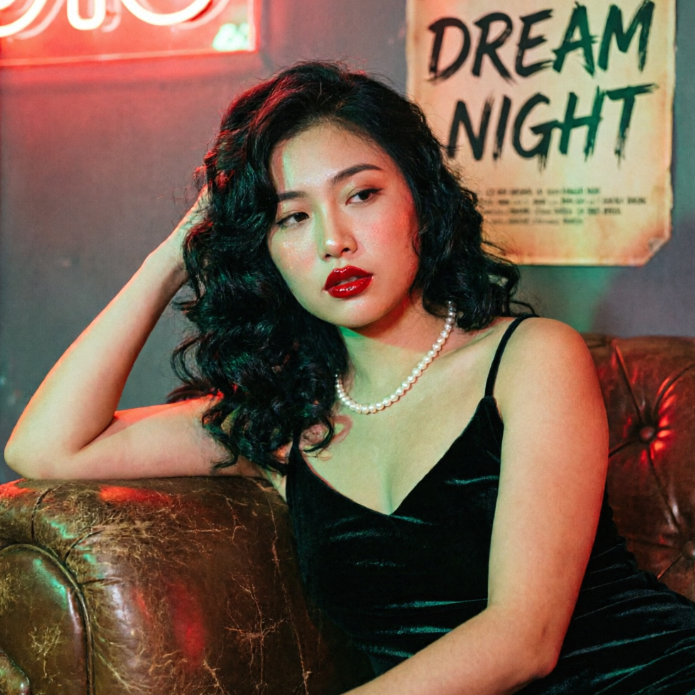 A young woman with black wavy hair sitting on a brown leather sofa, wearing a black velvet slip dress and a pearl necklace, with exquisite makeup and striking red lips. A vintage poster on the wall behind her reads "DREAM NIGHT" in green handwriting, complemented by red neon lighting to create a rich retro cinematic atmosphere.