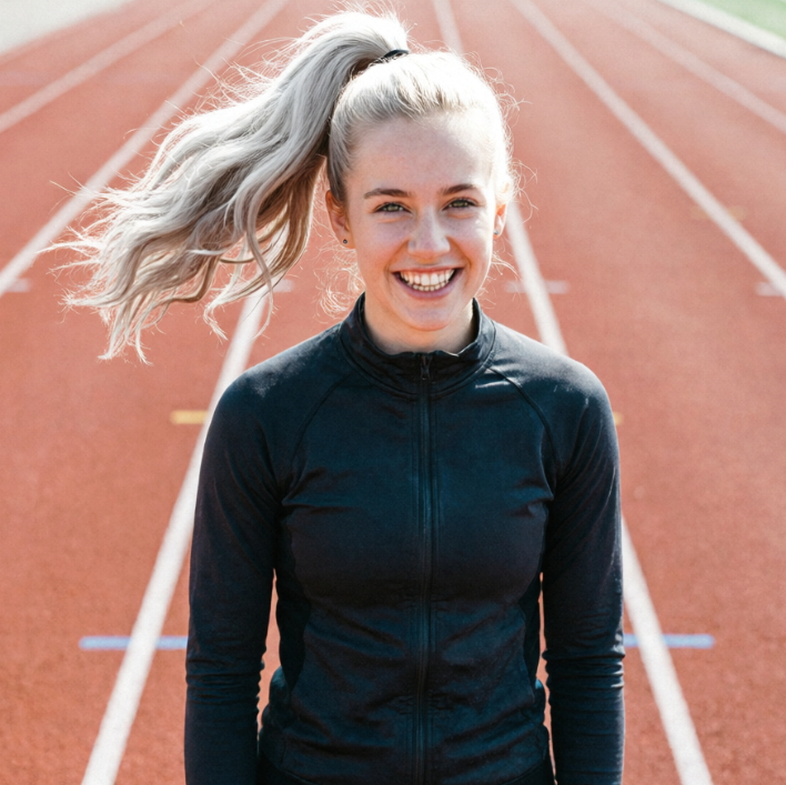 A young blonde woman with a high ponytail standing on a red running track, wearing a black athletic jacket and smiling brightly, full of energy.