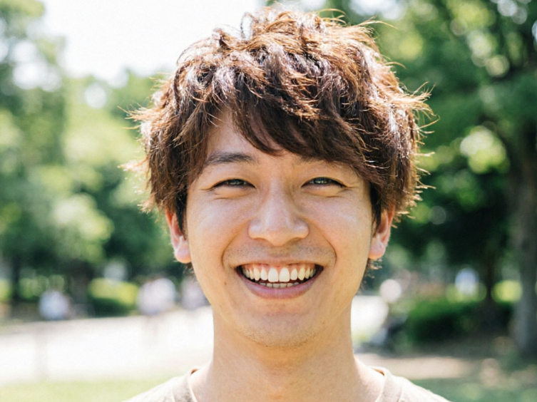 A young Asian man with messy short brown hair laughing heartily in a sunny park, with green trees in the background.