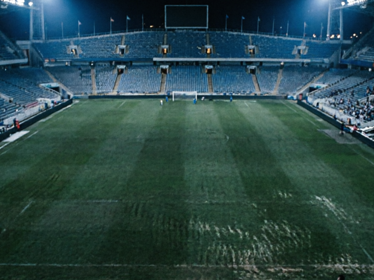 The original low-resolution version of the same football stadium. The image is dark and blurry overall, with indistinct grass textures, severe loss of spectator detail, dull colors, and lack of depth — typical of unprocessed source material.