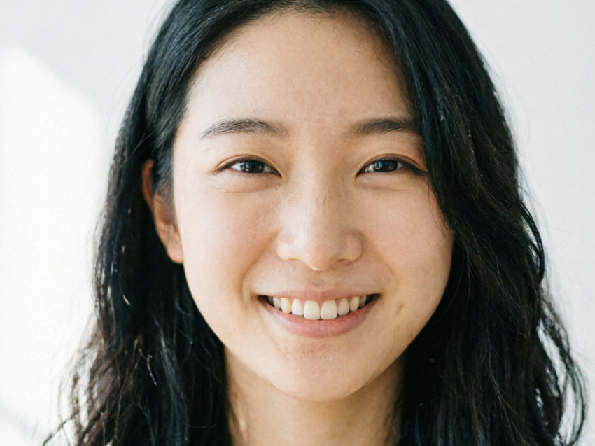 A close-up of a young Asian woman with long black wavy hair. She offers a sweet smile showing white teeth, with natural skin texture and bright eyes, against a simple white wall background.