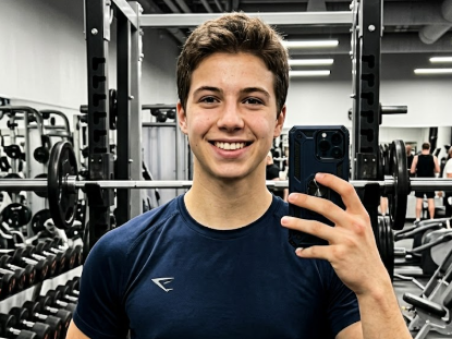 A young man takes a selfie in a gym, wearing a navy blue athletic T-shirt, smiling brightly. Background features dumbbell racks, barbells, and fitness equipment under bright lighting — energetic and confident.