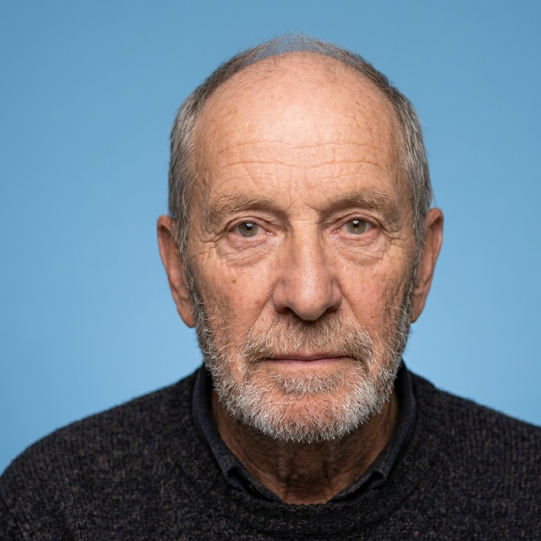 An elderly man with thinning gray hair and a graying beard looks straight ahead, dressed in a dark sweater against a light blue backdrop. His gentle yet profound expression reflects wisdom and serenity shaped by time.