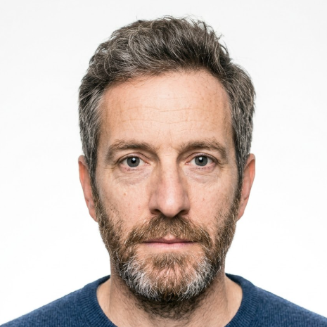 A middle-aged man with salt-and-pepper hair and beard gazes directly at the camera, wearing a navy blue knit sweater against a pure white background. His steady eyes and defined facial features convey maturity and composure.