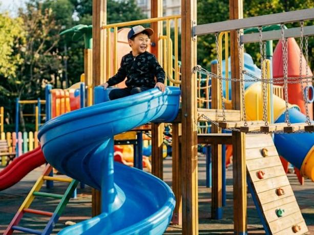The same young boy wearing a baseball cap sits atop a blue spiral slide in an outdoor playground, laughing happily in a dark camouflage shirt. The background features colorful play equipment, wooden structures, and green trees under bright sunlight, full of energy and childhood joy.
