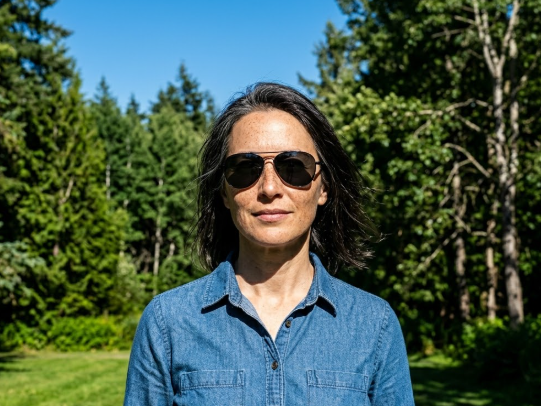 A middle-aged woman stands outdoors against a forest background on a sunny day. She wears aviator sunglasses and a blue denim shirt, with windblown hair and a relaxed demeanor.