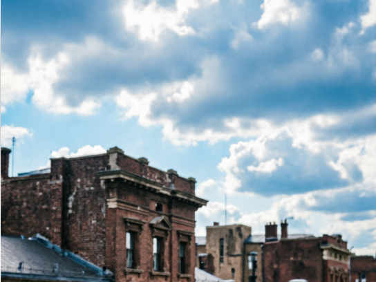 A cityscape featuring red brick buildings under a bright blue sky with fluffy white clouds. Architectural details like chimneys and windows are sharp, and the colors are saturated.