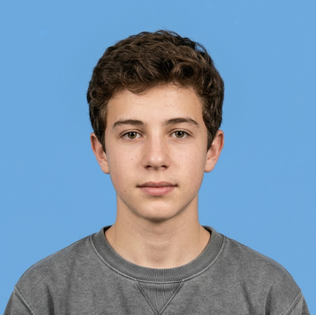 A young man with curly brown hair looks directly at the camera, wearing a gray crewneck sweatshirt against a solid blue background. His expression is calm and natural, evoking a clean, fresh portrait or ID photo style.