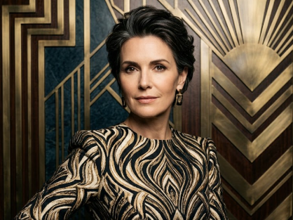 A middle-aged woman in a black-and-white wavy-patterned gown with gold earrings stands before an ornate geometric wall panel. Elegant lighting highlights her face — sophisticated and high-fashion.