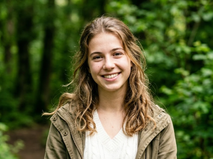 A young woman smiles in a lush forest path, her brown curly hair flowing naturally. She wears a beige knit top and olive green jacket, with soft natural light and blurred trees in the background — relaxed and joyful.