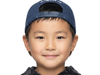 A frontal portrait of a young Asian boy against a solid white background. He is wearing a dark blue baseball cap backwards and a dark camouflage hoodie, smiling gently at the camera with even lighting, styled as a standard ID photo.