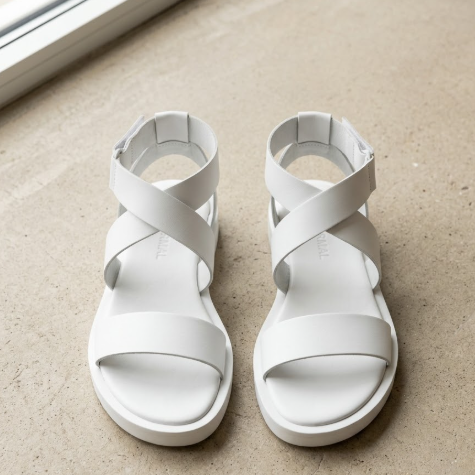 A pair of simply designed, streamlined white leather minimalist sandals, placed on a clean beige concrete background. The sandals are composed of just a few wide straps with no superfluous decoration, showcasing pure geometric aesthetics. Featuring a flat sole, slightly thick. The lighting is bright and even, highlighting the material and contours of the shoes. 英文简短Alt: AI designed white minimalist flat sandals.