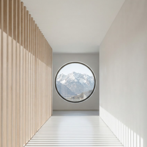 A 1:1 architectural study of a long corridor in a Scandinavian-style home. The left wall is made of vertical light wood slats, creating a rhythmic shadow pattern on the white floor. At the end of the hallway, a large circular window frames a mountain view.