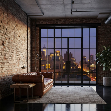 A 1:1 interior shot of an industrial loft living room. Exposed red brick walls contrast with polished black concrete floors. A large cognac leather sofa sits in the center