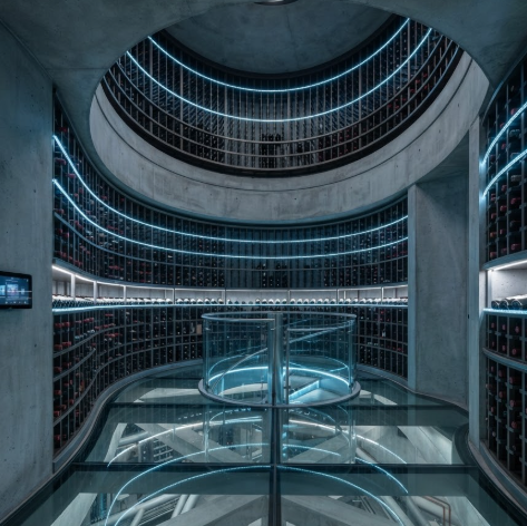 A 1:1 view of a high-tech underground wine cellar. Circular wine racks are built directly into curved concrete walls, illuminated by blue fiber-optic lights. The floor is tempered glass showing structural supports below.