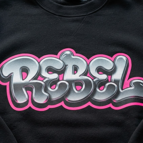 A close-up 1:1 shot of the chest area of a high-quality black cotton hoodie. A large, colorful graffiti logo is screen-printed on the fabric. The design shows "REBEL" in a 3D bubble style with a chrome-like metallic gradient and hot pink outlines. The fabric's knit texture is visible through the ink, showing high-end manufacturing quality.