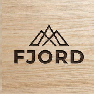 A 1:1 architectural shot. A logo for a furniture brand named "FJORD" laser-engraved onto a light ash wood surface. The design is stark and clean, featuring a minimalist mountain peak icon above the text. The wood grain is sharp and detailed. The style is Scandinavian, emphasizing craftsmanship and natural materials.