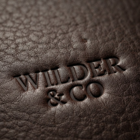 1:1 ratio. A deep brown, high-quality pebbled leather surface. The logo "WILDER & CO" is heat-embossed with significant depth, creating natural shadows within the indentations. The leather grain is hyper-realistic, showcasing extreme detail in the material's texture.