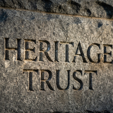 1:1 ratio. A dark grey granite building facade with the logo "HERITAGE TRUST" deeply carved into the stone. The carved grooves show natural weathering and aging effects. The morning sun hits the wall at an angle, creating long shadows that emphasize the 3D depth of the engraving.