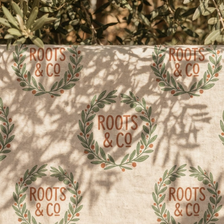 A 1:1 flat lay of an eco-conscious logo. The name "ROOTS & CO" is framed by an earthy, hand-illustrated olive branch wreath. The color palette is sage green and terra cotta. The logo is screen-printed on a raw linen tote bag. Natural sunlight filters through leaves, casting organic shadows across the fabric and the logo.