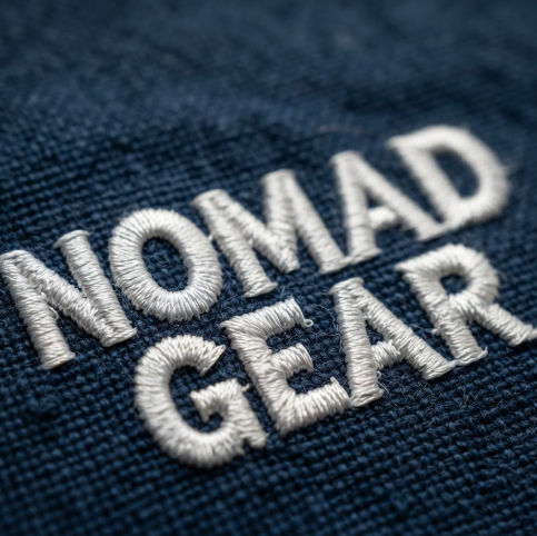 1:1 ratio. A close-up of a high-density white thread embroidery on navy blue canvas fabric. The logo "NOMAD GEAR" is depicted with visible stitching details, creating a 3D tactile effect. The background shows the coarse weave of the heavy-duty canvas material.