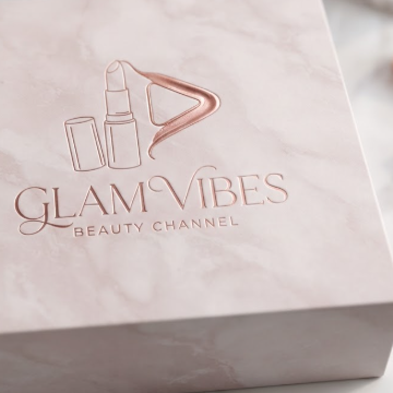 A 1:1 elegant YouTube logo for a beauty channel. A minimalist line-art drawing of a lipstick tube that leaves a trail in the shape of a rose-gold play button. Soft pink and marble textures. "GLAM VIBES" in elegant, thin cursive typography. The logo is embossed on a high-end luxury cosmetics box.